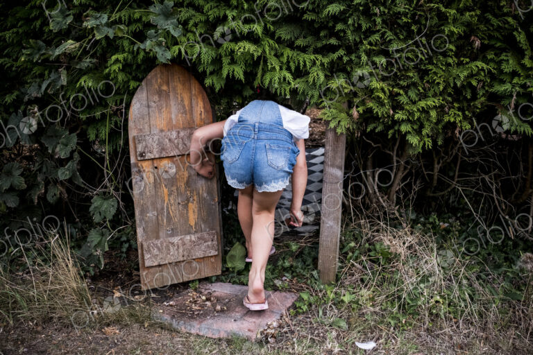 Rear view of girl wearing denim dungarees walking through small wooden door in a…