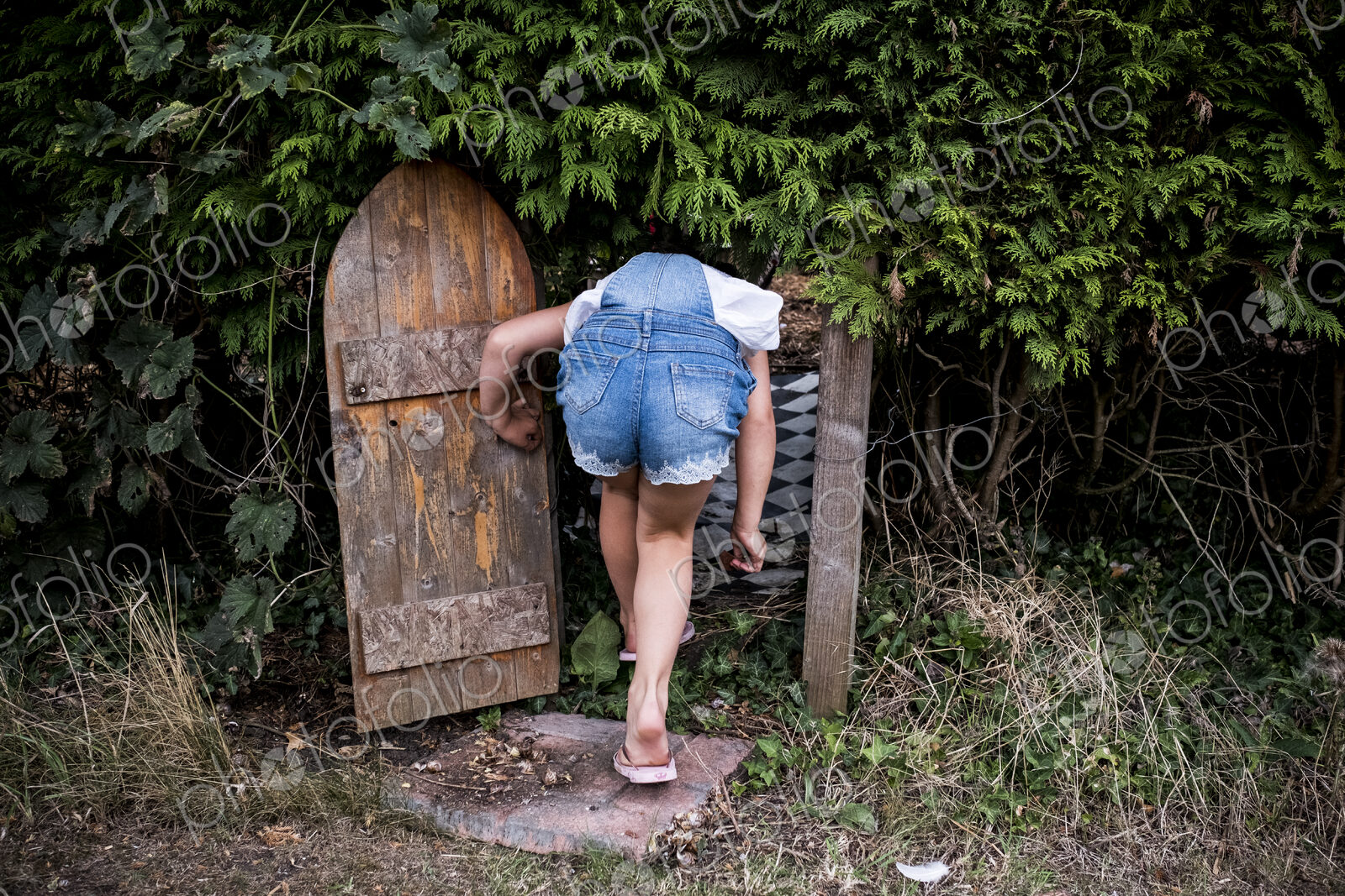 Rear view of girl wearing denim dungarees walking through small wooden door in a…