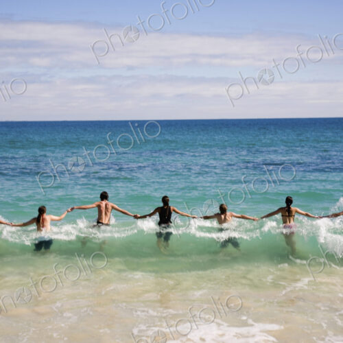 Rear view of group of people holding hands, jumping into wave in ocean.