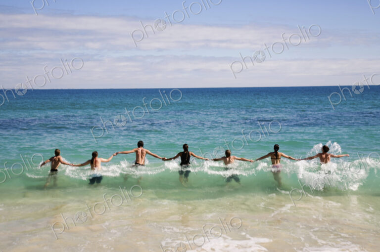 Rear view of group of people holding hands, jumping into wave in ocean.