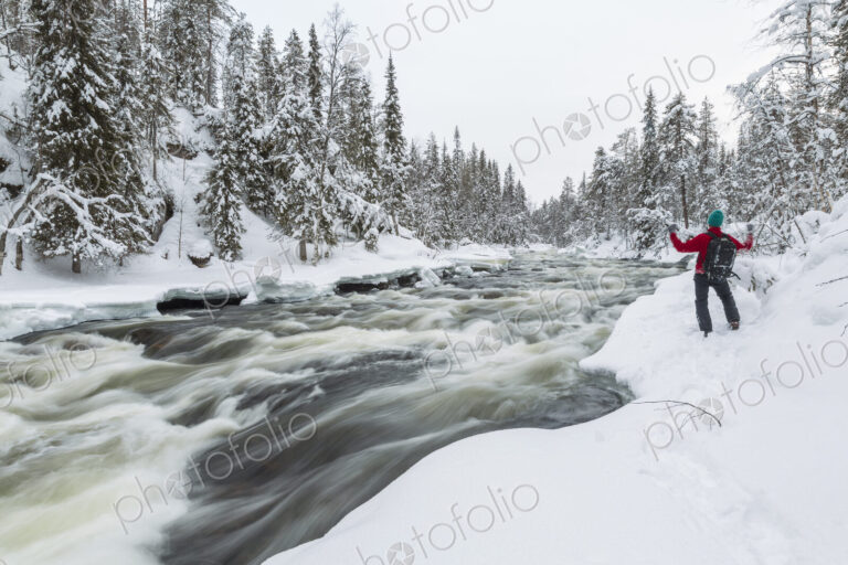Rear view of man standing by rapid in forest with snow-covered trees.