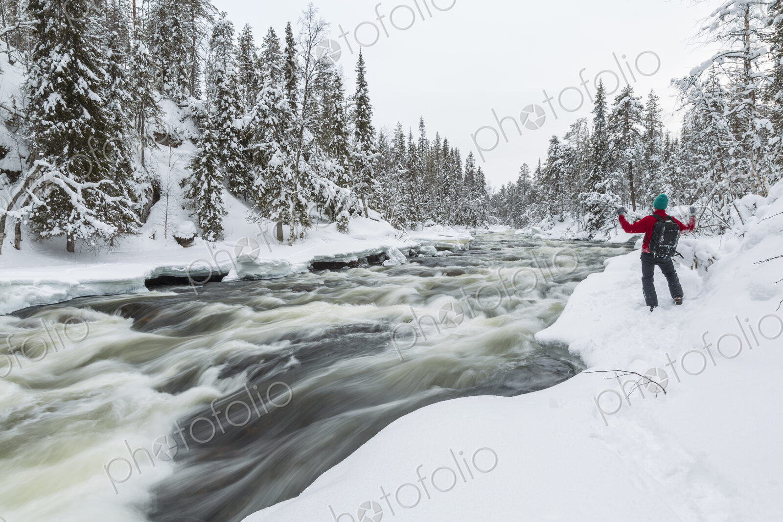 Rear view of man standing by rapid in forest with snow-covered trees.