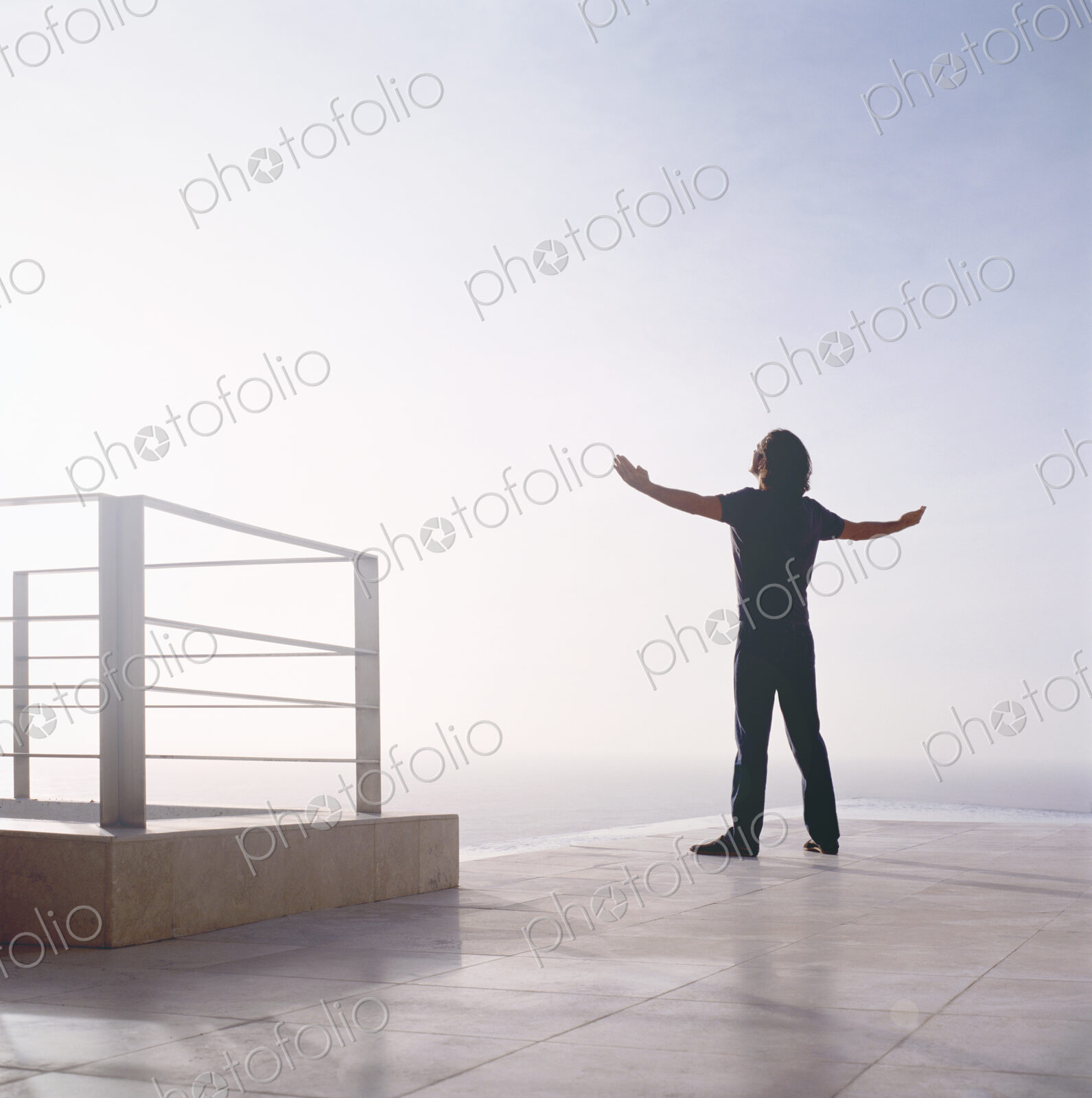 Rear view of man standing outdoors at edge of the ocean, arms outstretched.