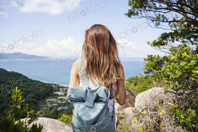 Rear view of woman carrying backpack standing on a rock on cliff, ocean in the background.