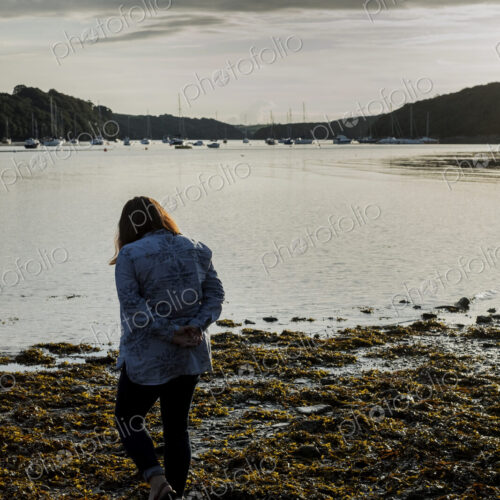 Rear view of woman standing on a beach at sunset, with sailboats and cliffs in the… Rear view of woman standing on a beach at sunset, with sailboats and cliffs in the…
