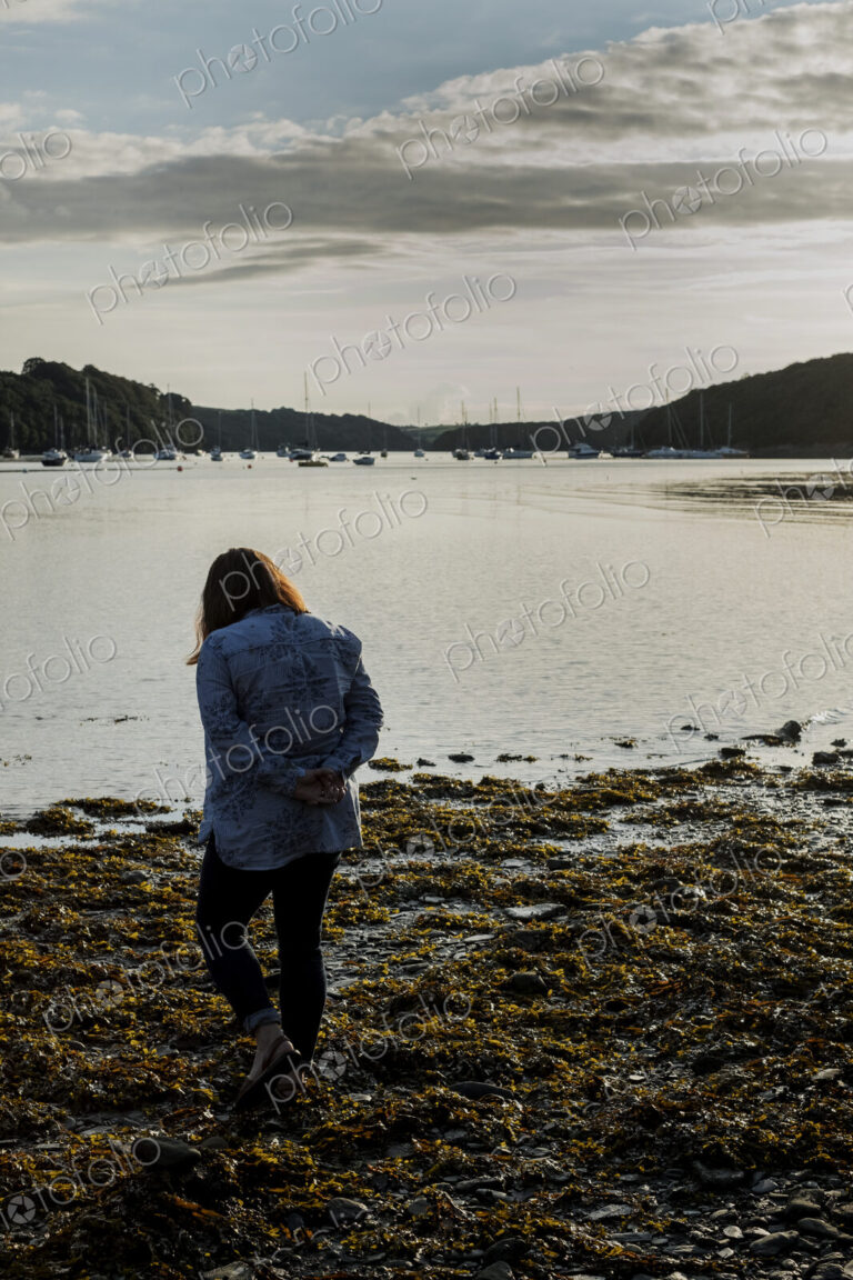 Rear view of woman standing on a beach at sunset, with sailboats and cliffs in the…