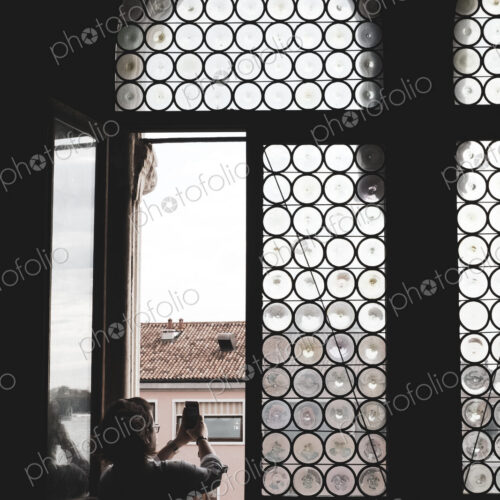 Rear view of woman taking picture through arched window with leaded glass in Venice,… Rear view of woman taking picture through arched window with leaded glass in Venice,…
