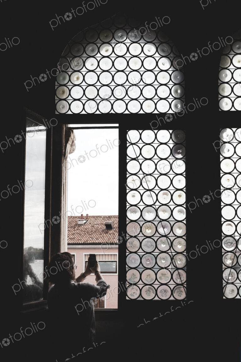 Rear view of woman taking picture through arched window with leaded glass in Venice,…