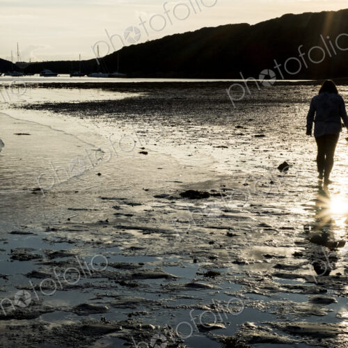 Rear view of woman walking across a sandy beach at sunset, with sailboats and cliffs… Rear view of woman walking across a sandy beach at sunset, with sailboats and cliffs…