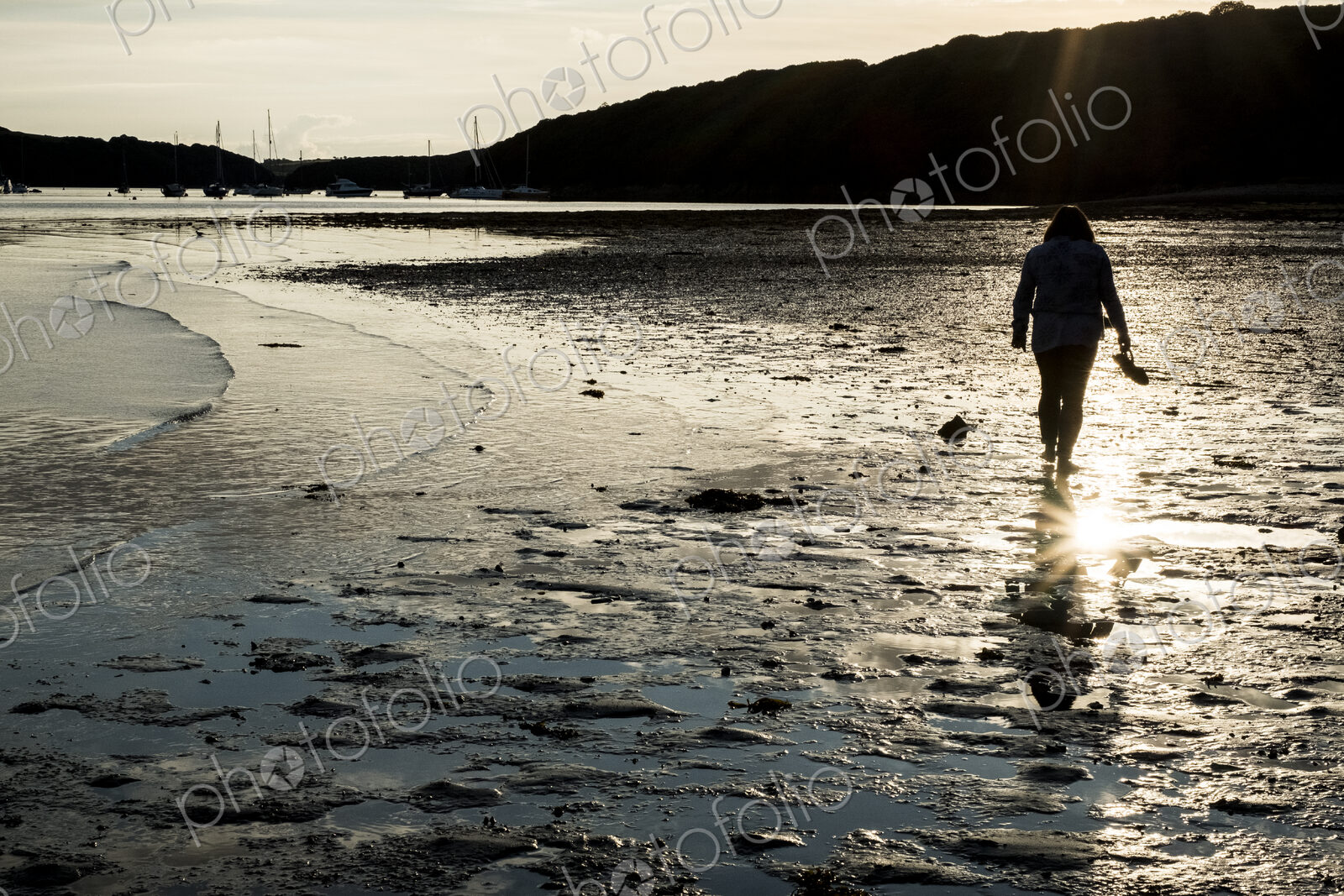 Rear view of woman walking across a sandy beach at sunset, with sailboats and cliffs…