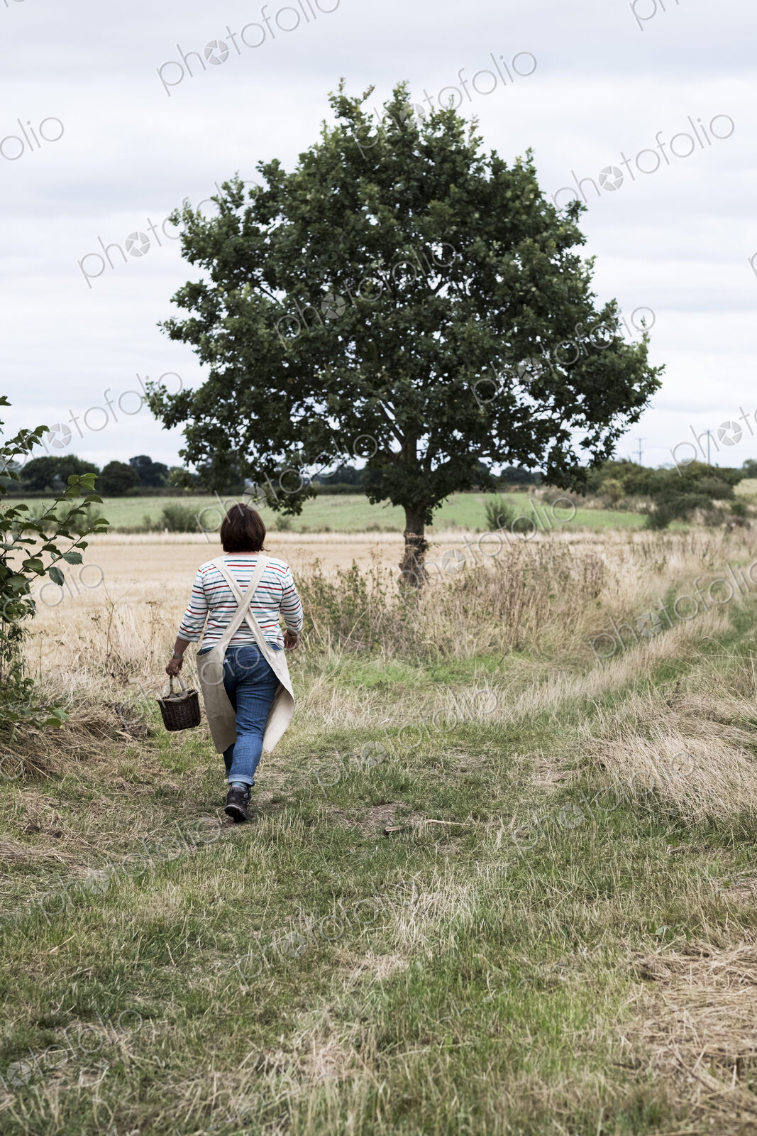 Rear view of woman walking along rural path, carrying brown wicker basket.
