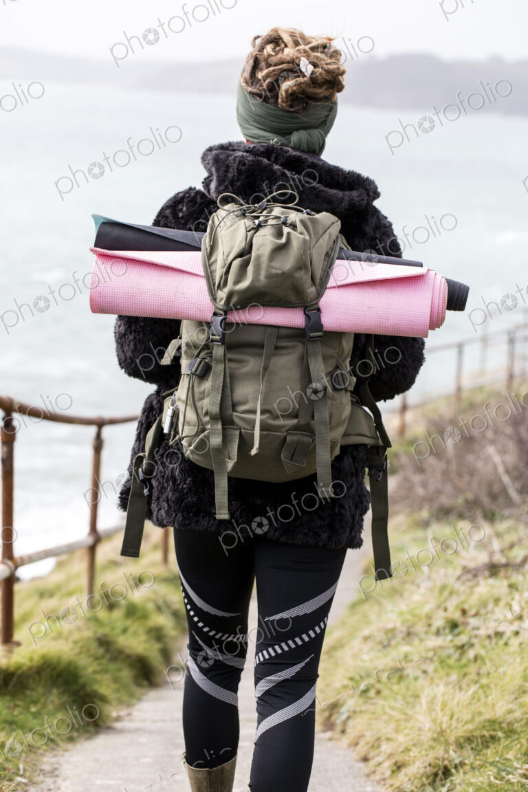Rear view of young woman with brown hair and dreadlocks carrying backpack, walking…