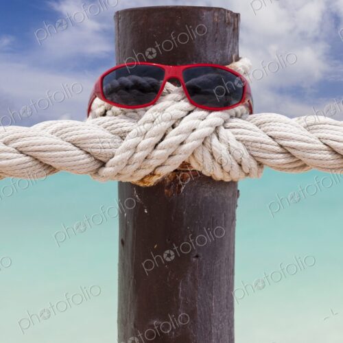Red sunglasses rest on a wooden post wrapped in nautical rope, with turquoise…