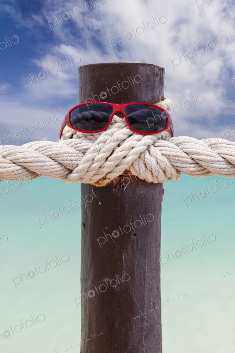 Red sunglasses rest on a wooden post wrapped in nautical rope, with turquoise…