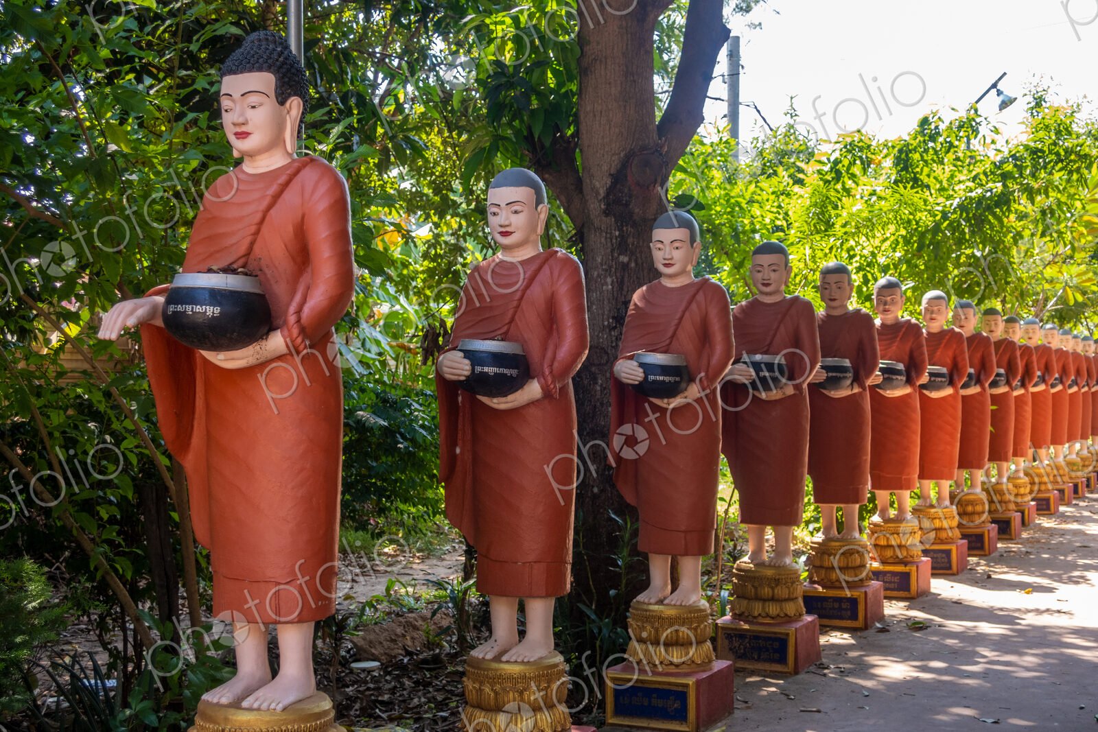 Row of Buddhist monk statues with red robes and alms bowls in the gardens of the…