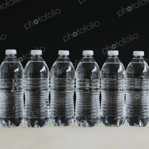Row of clear, plastic water bottles filled with filtered water in a row, on a black…