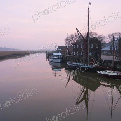 Sailing boats moored at harbour wall, reflected on the surface of a calm canal at… Sailing boats moored at harbour wall, reflected on the surface of a calm canal at…