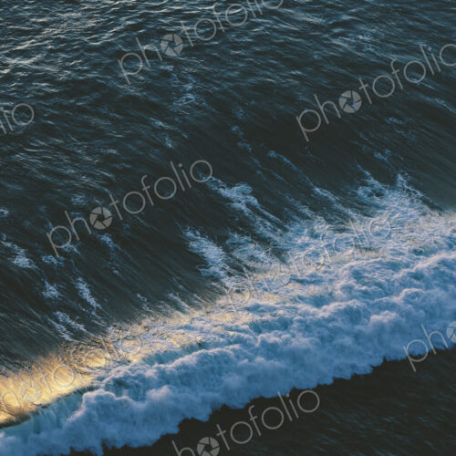 Seascape abstract at dusk, surf breaking on the shore, overhead view and sunlight on…