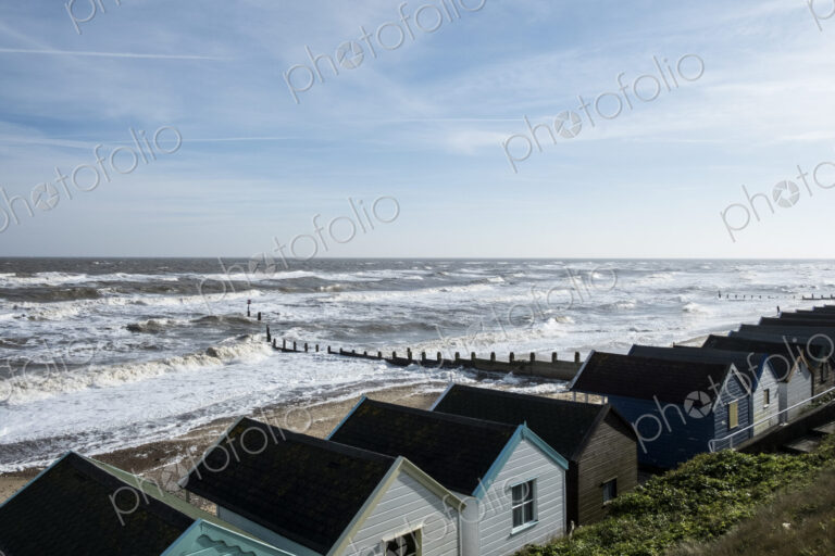 Seascape over a row of wooden beach huts, with waves rolling onto beach near groyne.