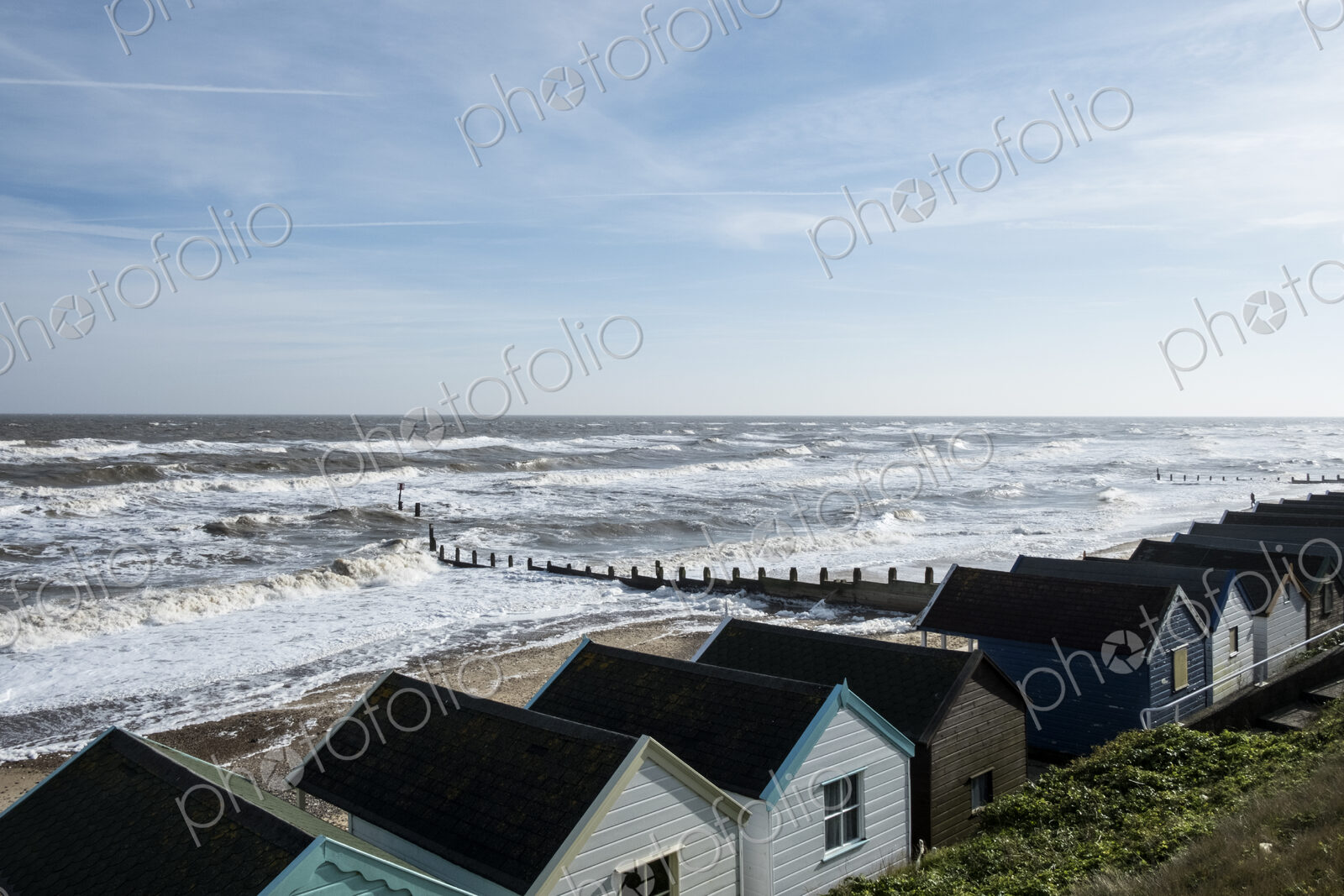 Seascape over a row of wooden beach huts, with waves rolling onto beach near groyne.