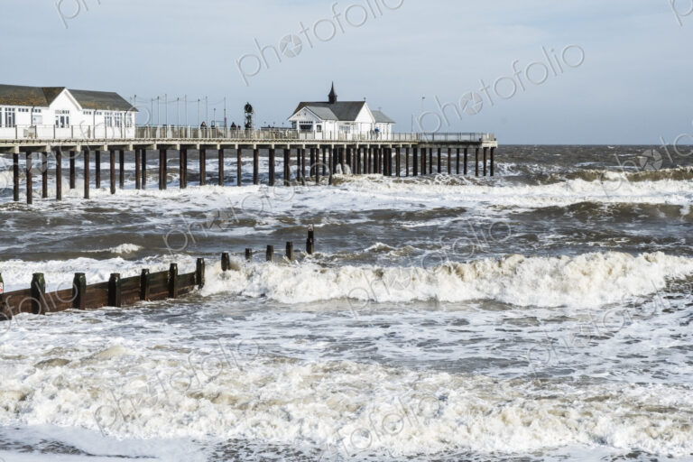Seascape with waves rolling onto beach near groyne and white wooden buildings on a pier.