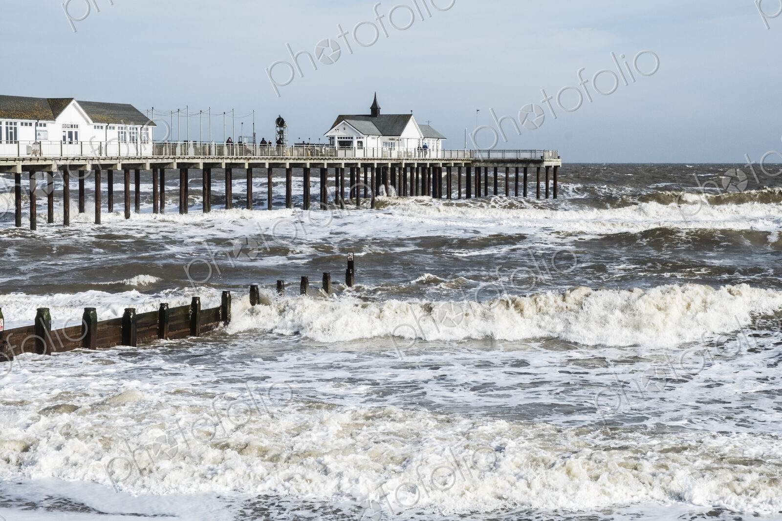 Seascape with waves rolling onto beach near groyne and white wooden buildings on a pier.