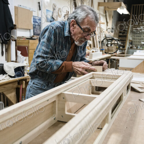 Senior Caucasian carepenter sanding a wooden cabinet part in a large woodworking shop.