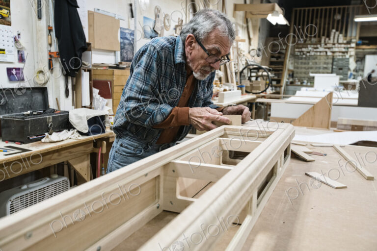 Senior Caucasian carepenter sanding a wooden cabinet part in a large woodworking shop.
