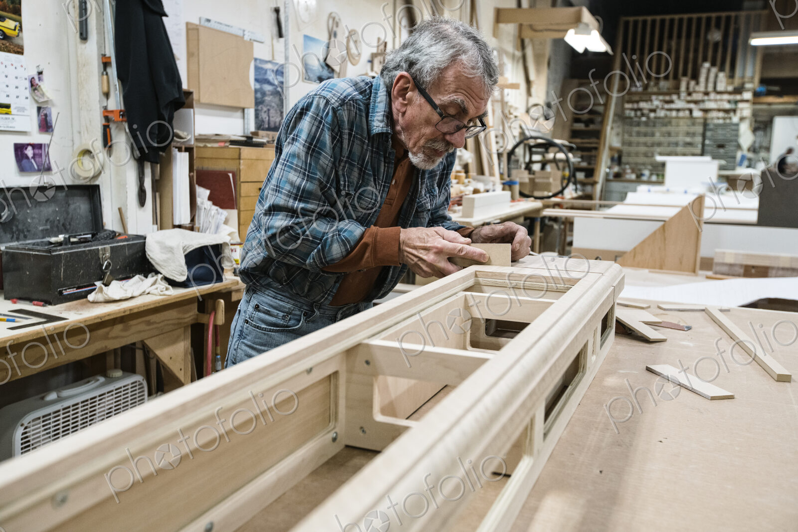 Senior Caucasian carepenter sanding a wooden cabinet part in a large woodworking shop.