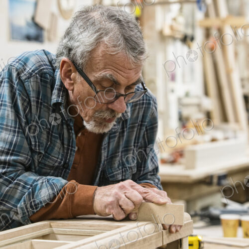 Senior Caucasian carepenter sanding a wooden cabinet part in a large woodworking shop.