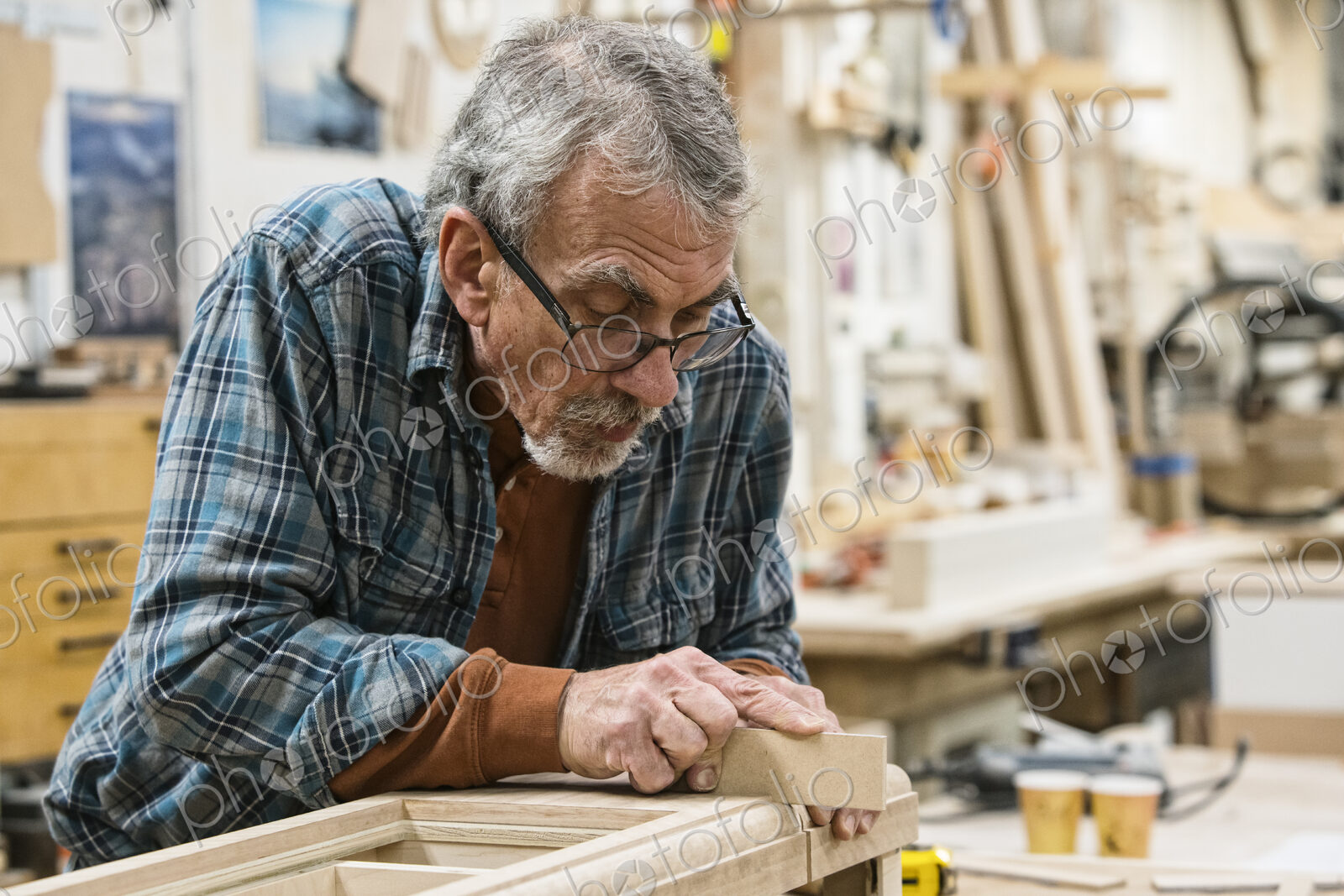 Senior Caucasian carepenter sanding a wooden cabinet part in a large woodworking shop.