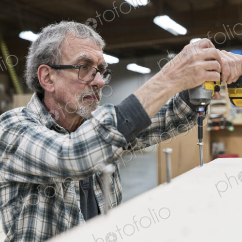 Senior carpenter drilling a screw into a cabinet in a large woodworking factory. Senior carpenter drilling a screw into a cabinet in a large woodworking factory.