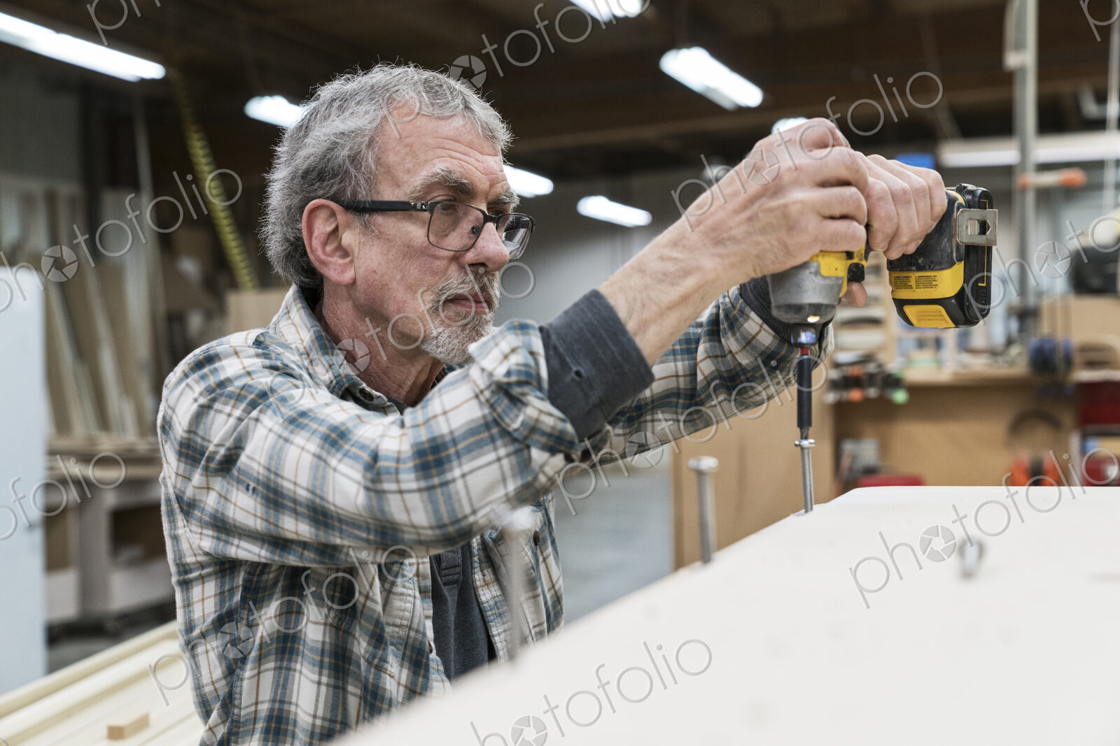 Senior carpenter drilling a screw into a cabinet in a large woodworking factory.