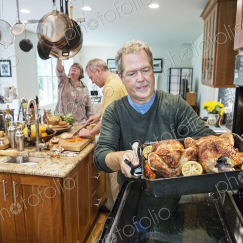 Senior man pull roast chickens out of the oven at a home dinner party.