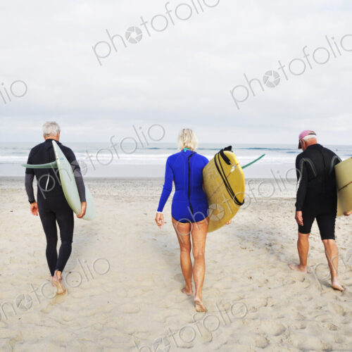 Senior woman and two senior men on a beach, wearing wetsuits and carrying surfboards. Senior woman and two senior men on a beach, wearing wetsuits and carrying surfboards.