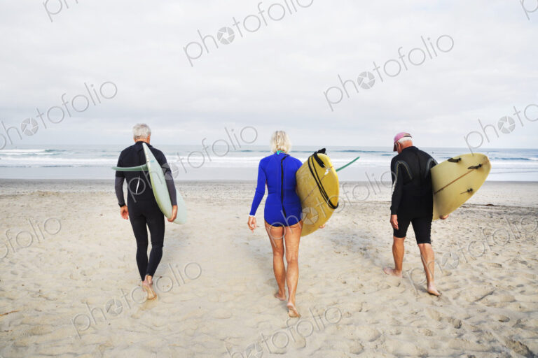 Senior woman and two senior men on a beach, wearing wetsuits and carrying surfboards.