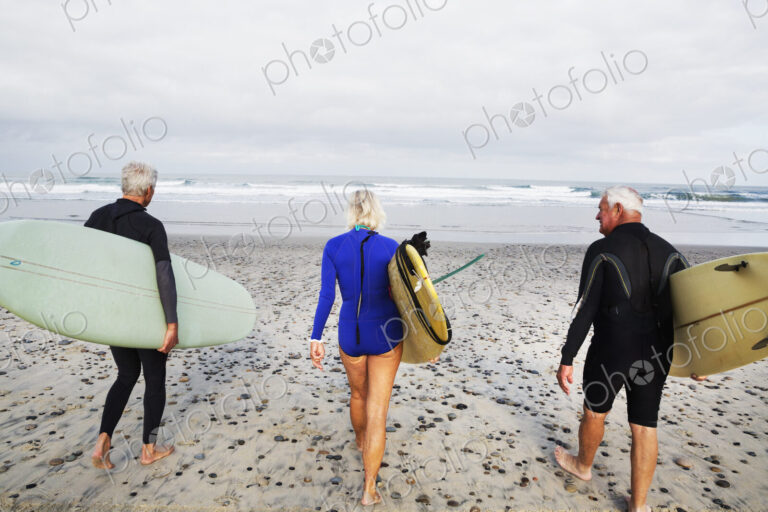 Senior woman and two senior men on a beach, wearing wetsuits and carrying surfboards.