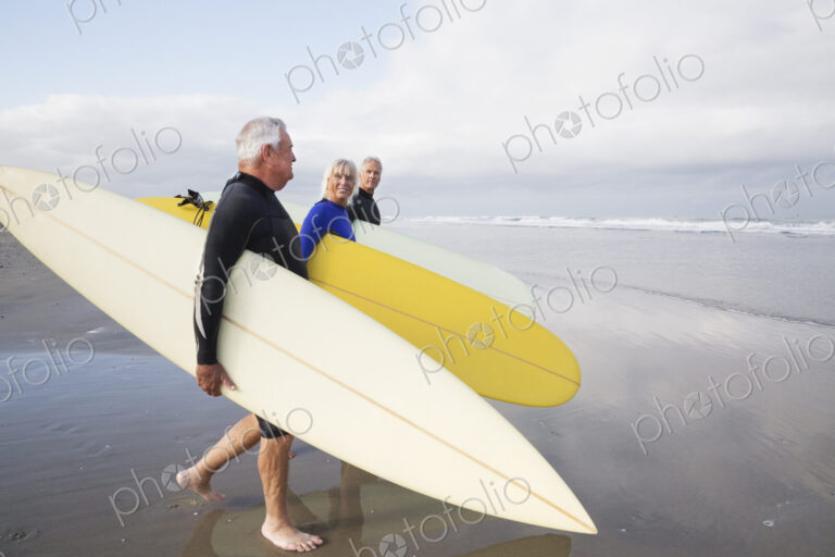 Senior woman and two senior men on a beach, wearing wetsuits and carrying surfboards.
