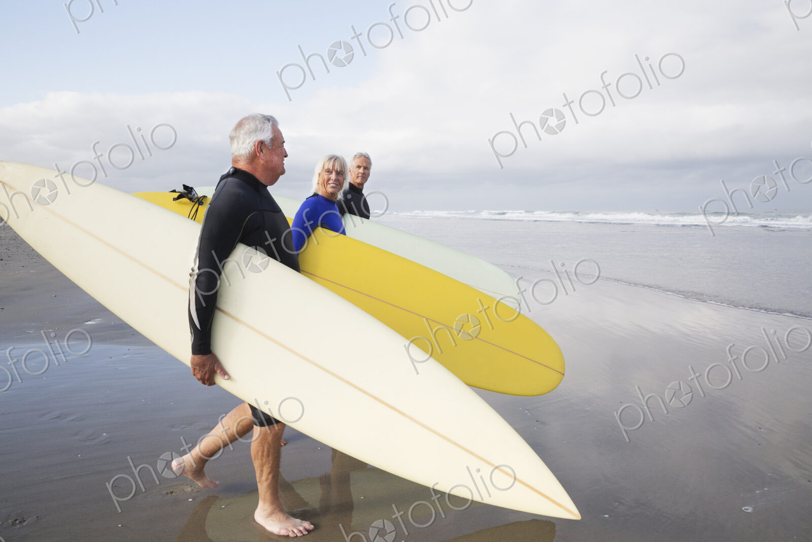 Senior woman and two senior men on a beach, wearing wetsuits and carrying surfboards.