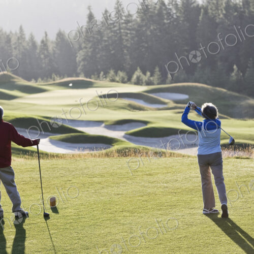 Senior woman golfer tees off with her male partner observing off to the side.