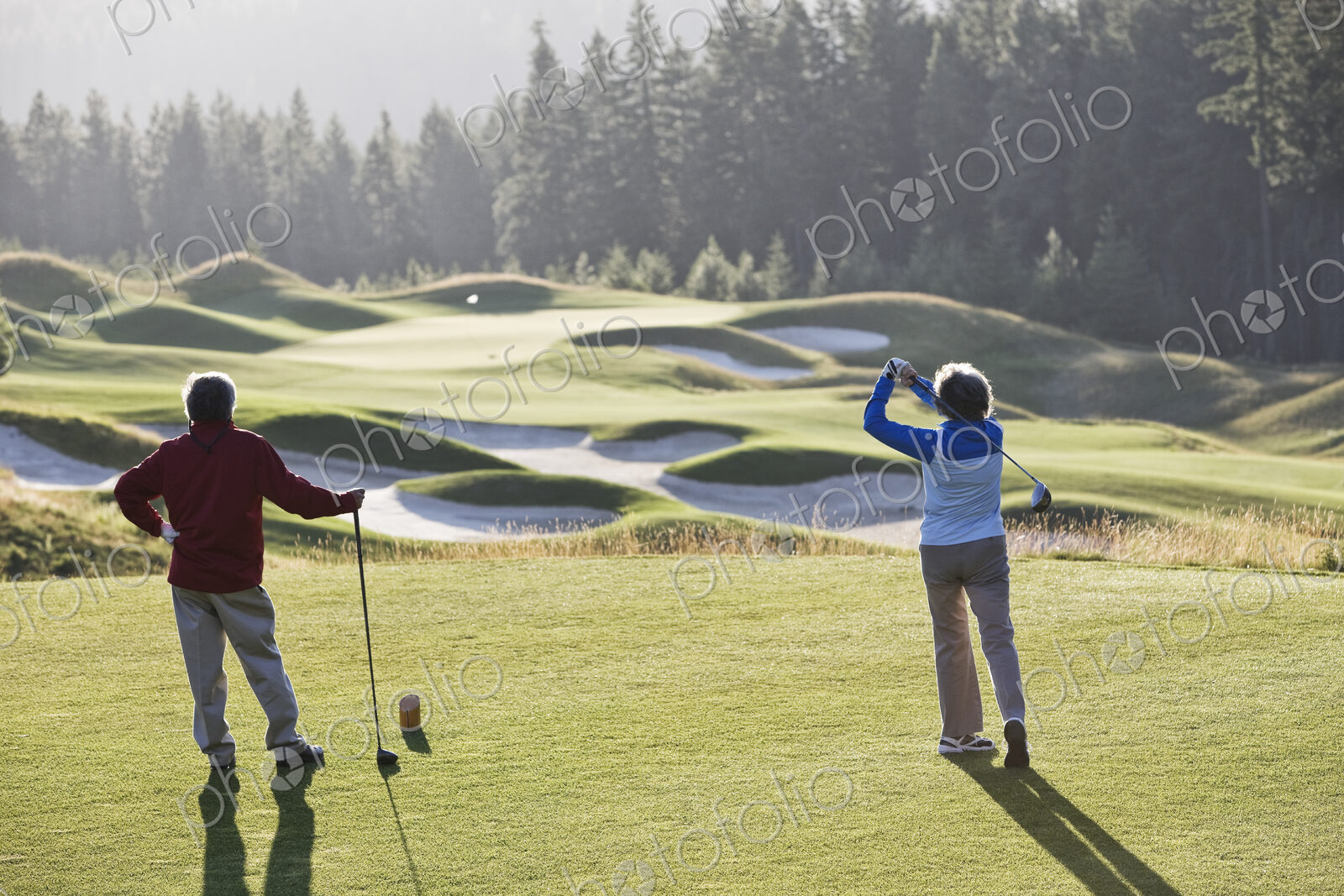 Senior woman golfer tees off with her male partner observing off to the side.