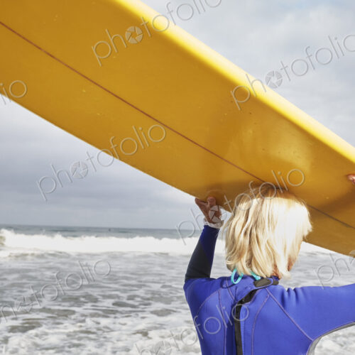 Senior woman on a beach, wearing a wetsuit and carrying a surfboard on her head. Senior woman on a beach, wearing a wetsuit and carrying a surfboard on her head.