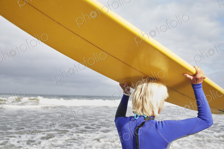 Senior woman on a beach, wearing a wetsuit and carrying a surfboard on her head.