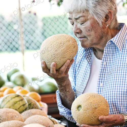 Senior woman standing at a farm stall, holding a fresh melon to her nose, smelling it. Senior woman standing at a farm stall, holding a fresh melon to her nose, smelling it.