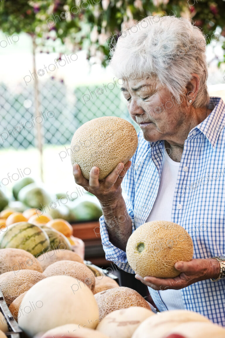 Senior woman standing at a farm stall, holding a fresh melon to her nose, smelling it.