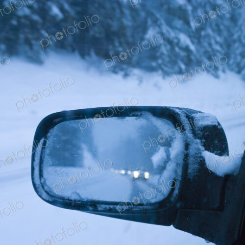 Side mirror reflection of snowy road at dusk and car headlights behind. Side mirror reflection of snowy road at dusk and car headlights behind.