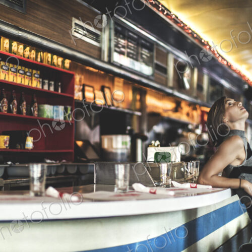 Side view of woman wearing jeans and sleeveless top leaning against a bar counter in a… Side view of woman wearing jeans and sleeveless top leaning against a bar counter in a…