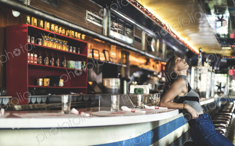 Side view of woman wearing jeans and sleeveless top leaning against a bar counter in a…