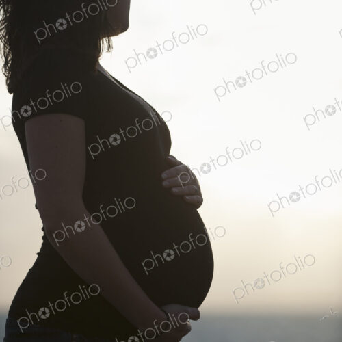 Silhouette of a heavily pregnant woman standing on a beach, with one hand resting on…