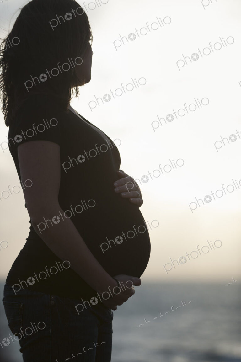 Silhouette of a heavily pregnant woman standing on a beach, with one hand resting on…